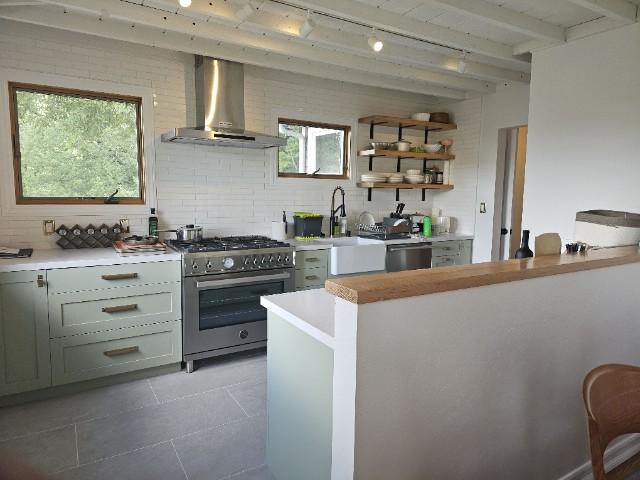 A modern kitchen with light green cabinets, a stainless steel stove and hood, wall-mounted shelves with dishes, a farmhouse sink, and a wooden countertop on a partial divider. Windows offer outdoor views.