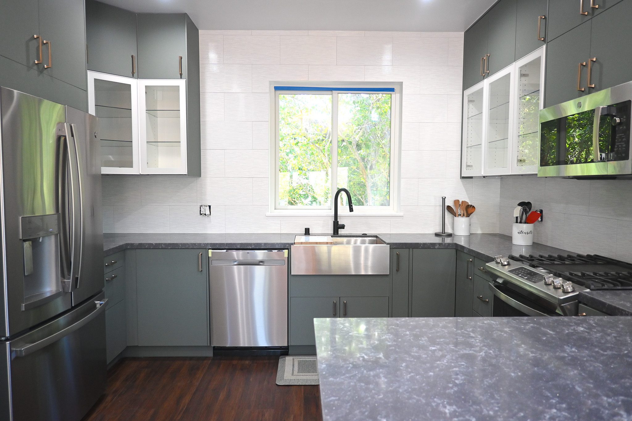 Modern kitchen with stainless steel appliances, grey cabinets, marble countertops, and a window above the sink. It features a large fridge, dishwasher, gas stove, and dark wood flooring.