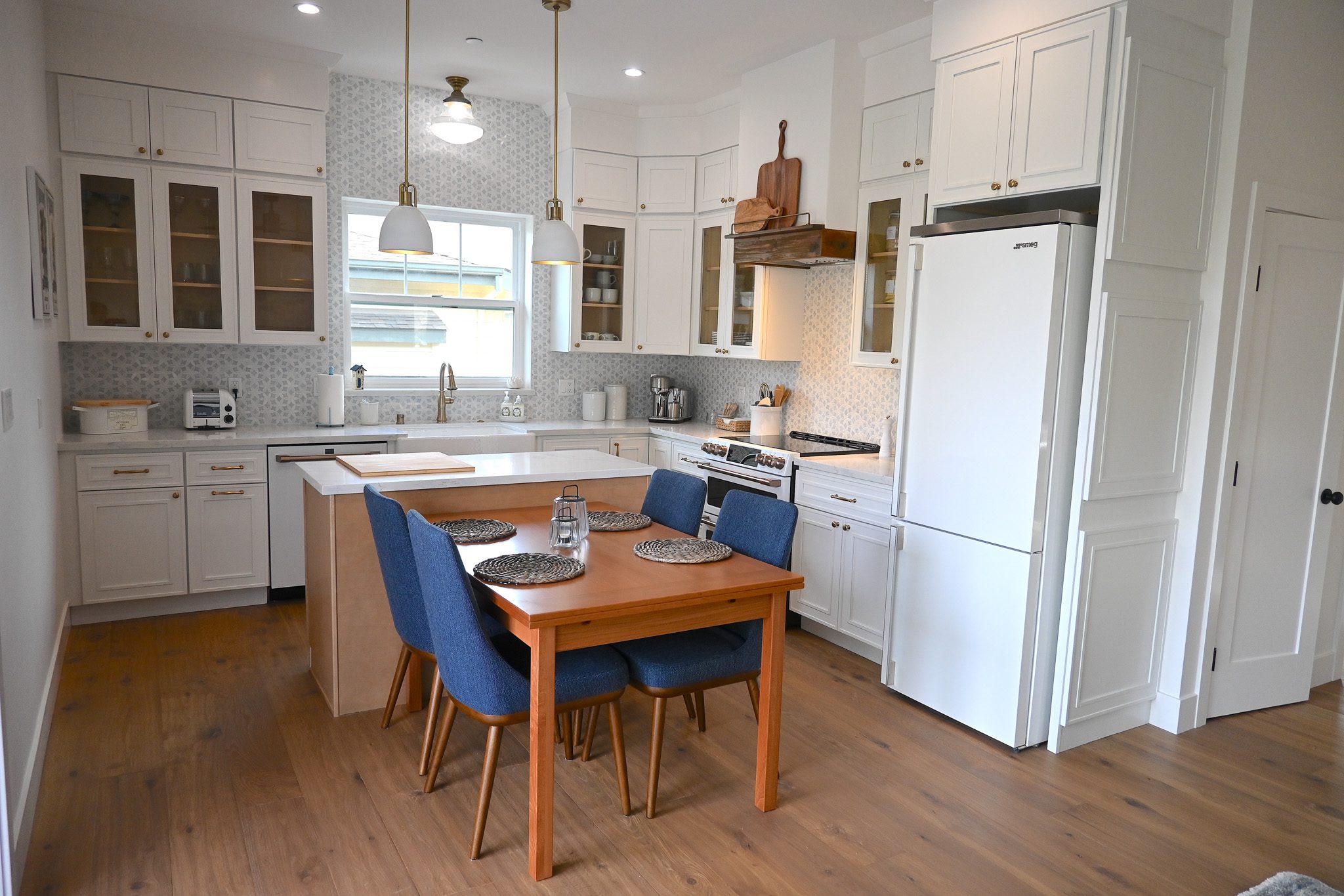 Modern kitchen with white cabinetry, stainless steel appliances, and a wooden dining table with four blue chairs. The kitchen island features a light wood finish and the room is well-lit by pendant lights.