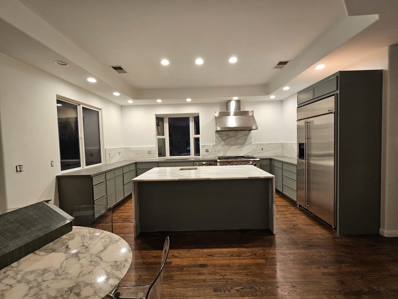 Modern kitchen with white cabinetry, stainless steel appliances, and a wooden dining table with four blue chairs. The kitchen island features a light wood finish and the room is well-lit by pendant lights.