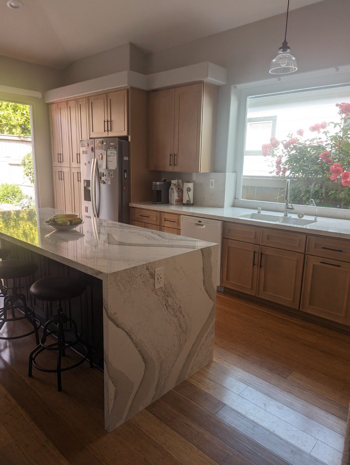 A modern kitchen with wooden cabinets, marble countertops, an island with seating, stainless steel refrigerator, and a large window with a view of flowering bushes.