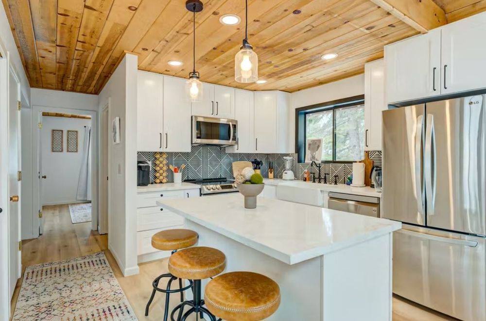 Modern kitchen with wooden ceiling, white cabinets, stainless steel appliances, and a central island with three brown bar stools. A large window and pendant lights provide ample illumination.