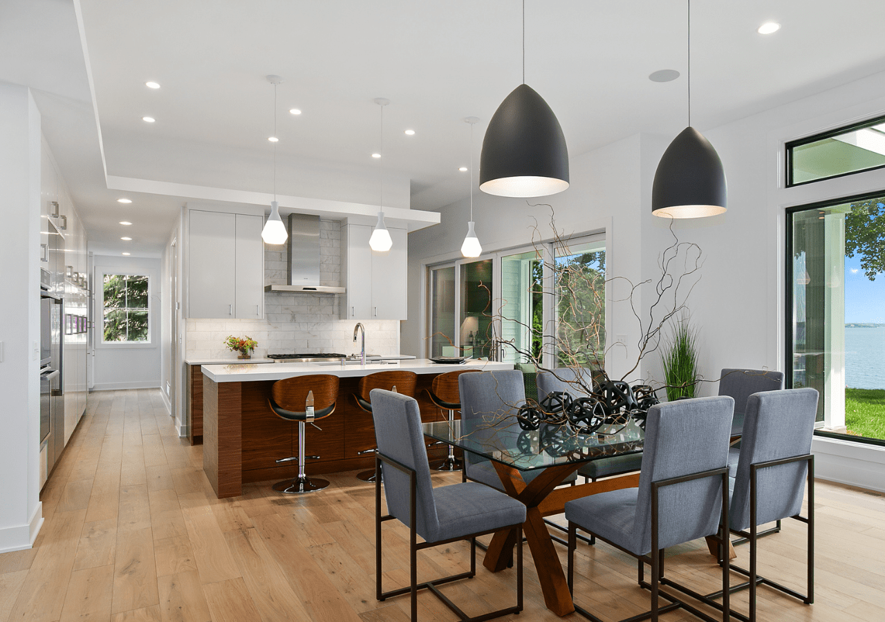 Modern kitchen and dining area with wooden floors, gray padded chairs, glass dining table, pendant lights, stainless steel appliances, and large windows showcasing a lake view.