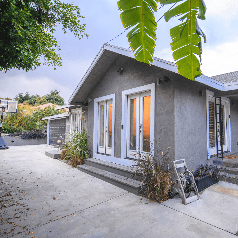 A modern single-story house with gray exterior, two French doors, and a concrete driveway. A basketball hoop is visible in the background. Lush plants surround the structure, with large leaves in the foreground.
