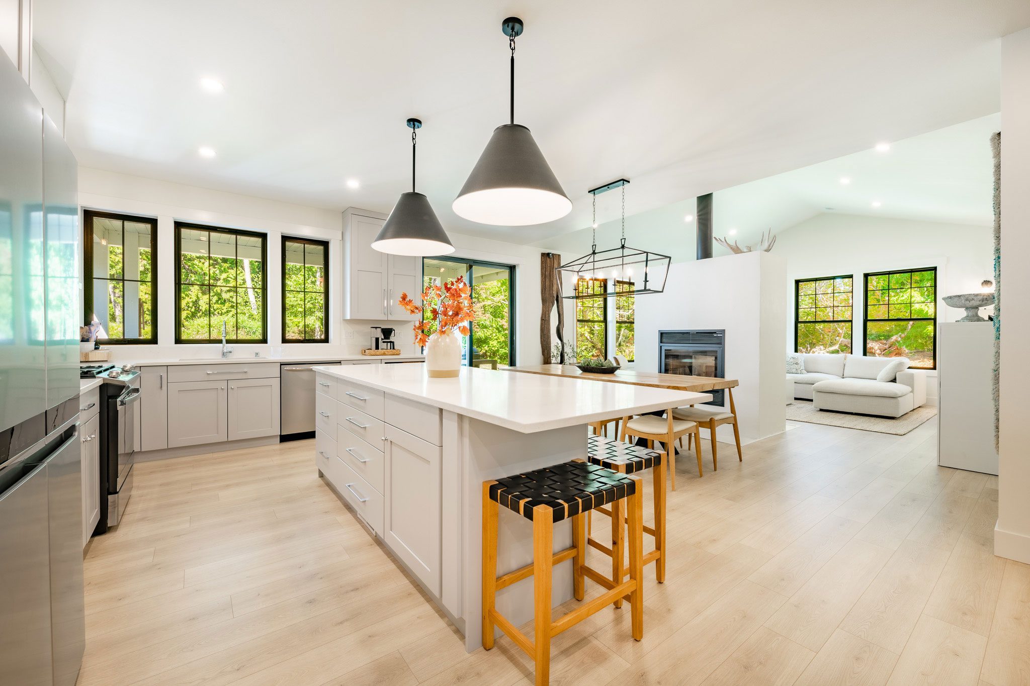 Modern kitchen with large island, pendant lights, and bar stools. In the background, there's a living area with a white sofa and large windows showcasing a green outdoor view.