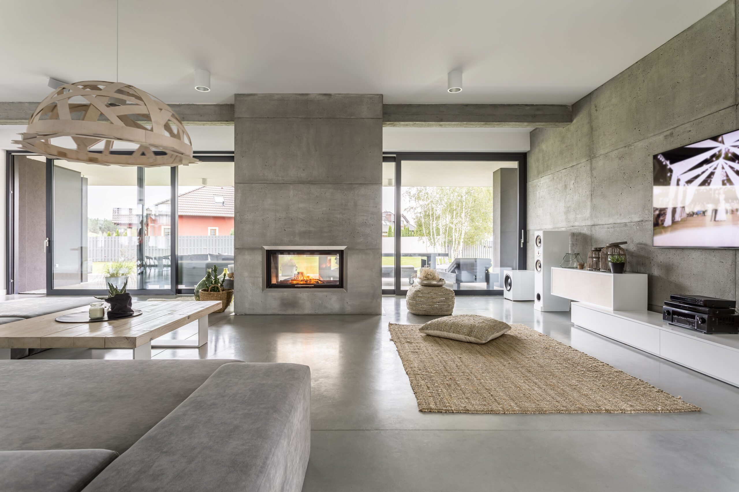 A modern living room with a concrete fireplace, large windows, a grey sofa, a wooden coffee table, a woven rug, and a mounted TV. The room has a minimalist design with ample natural light.