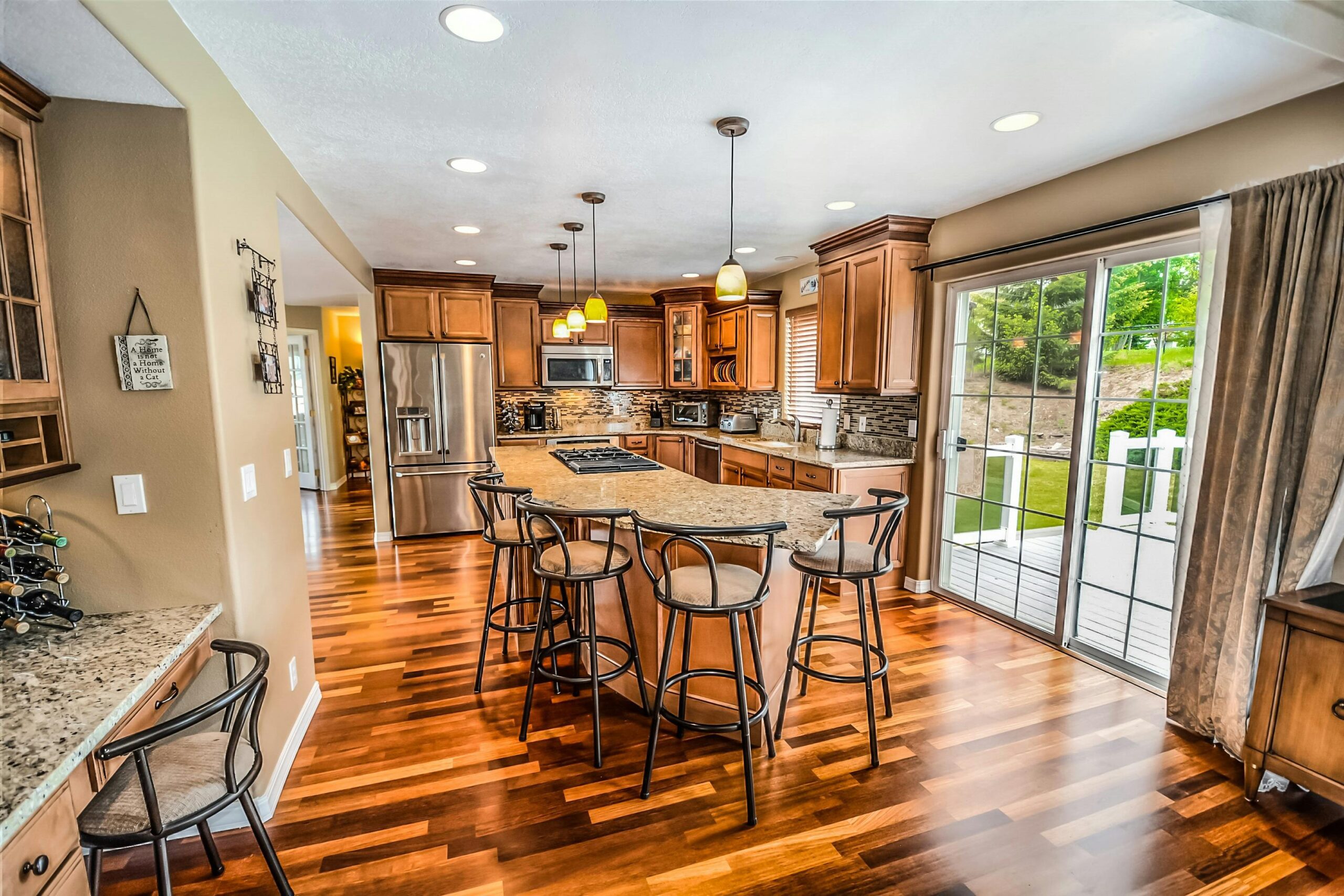 Modern kitchen with wooden cabinets, island seating, stainless steel appliances, pendant lights, and hardwood floors, adjacent to a glass sliding door leading to a deck and outdoor area.