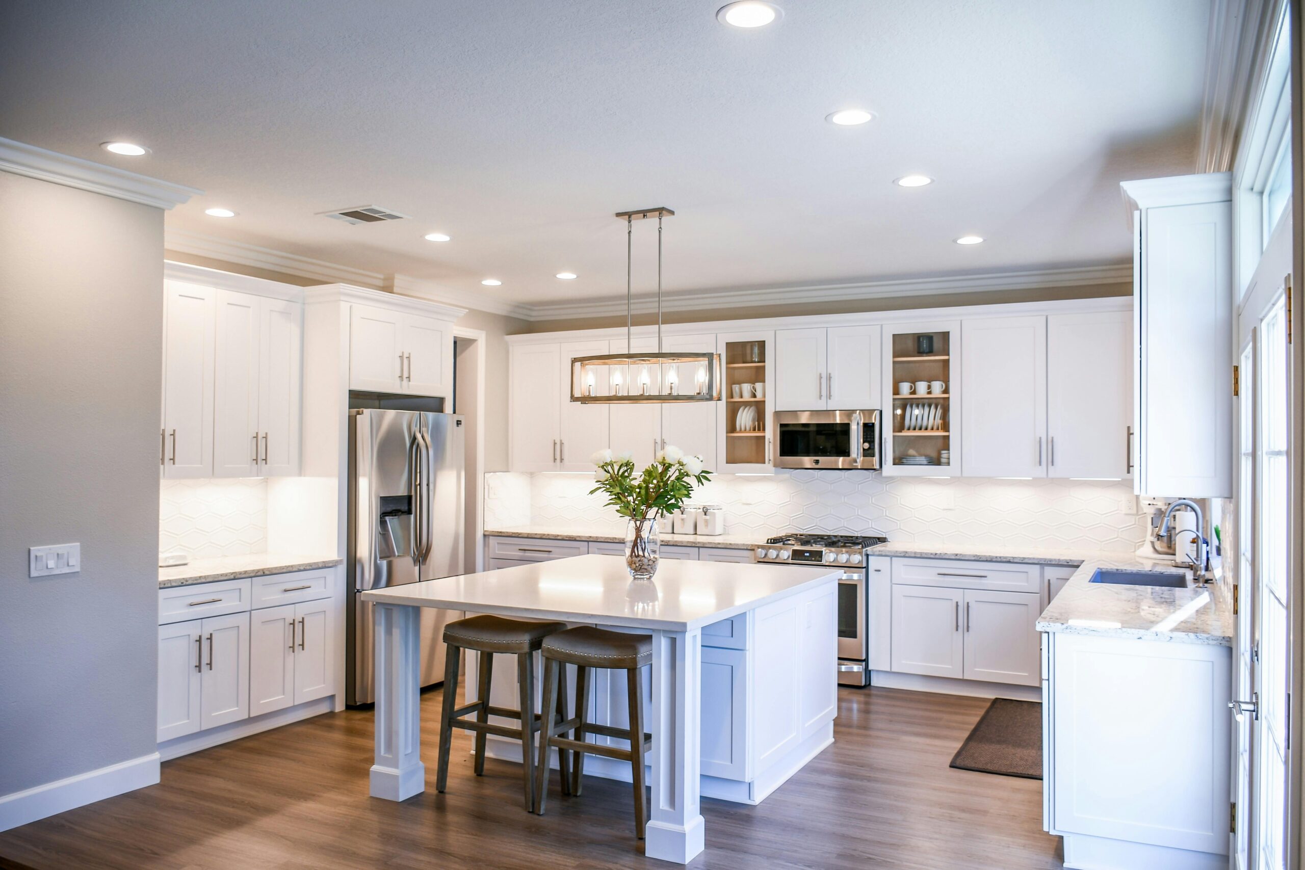 Modern kitchen with large island, pendant lights, and bar stools. In the background, there's a living area with a white sofa and large windows showcasing a green outdoor view.